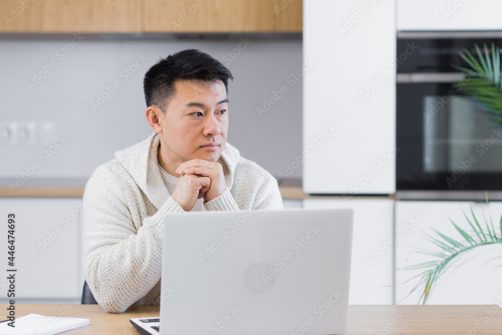 Thoughtful serious young asian man looking away. student writer sit at home office desk with laptop thinking of inspiration search solution ideas lost in thoughts dreaming solving financial problems Thoughtful serious young asian man looking away. student writer sit at home office desk with laptop thinking of inspiration search solution ideas lost in thoughts dreaming solving financial problems