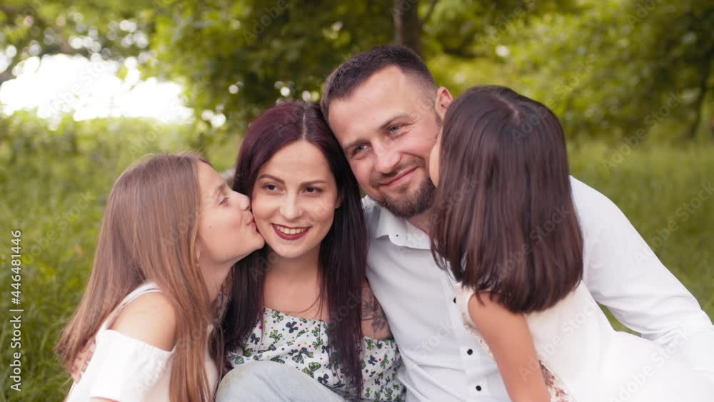 Two pretty sisters kissing their father and mother during summer picnic at green garden. Beautiful caucasian family relaxing together on fresh air. Love and happiness concept. Girls kissing Parents Two pretty sisters kissing their father and mother during summer picnic at green garden. Beautiful caucasian family relaxing together on fresh air. Love and happiness concept. Girls kissing Parents