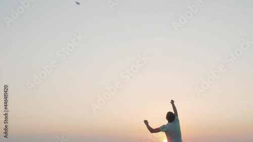 man in cheerful mood throwing paper airplane in field on sunset on background of clear sky, concept of active rest outdoors and relaxation, toy of childhood photo