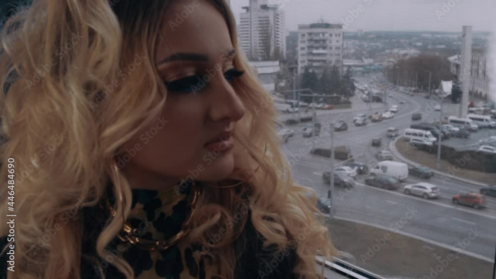 The young blonde, with curls and shrill make-up, looks to one side on the window at the view of the city center. Female energy. Beauty industry. Artistic concept The young blonde, with curls and shrill make-up, looks to one side on the window at the view of the city center. Female energy. Beauty industry. Artistic concept