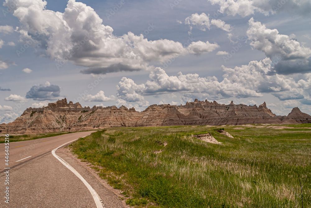 Badlands National Park, SD, USA - June 1, 2008: Road on side of green prairie in front of brown mountain range of geological deposits under spectacular light blue cloudscape.