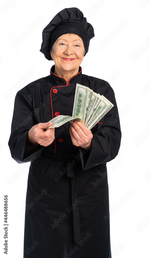 woman chef in black uniform. apron, shirt, hat. An elderly woman holds money, dollars fanned out. white background isolated woman chef in black uniform. apron, shirt, hat. An elderly woman holds money, dollars fanned out. white background isolated