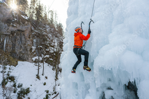 Aerial shot of an ice climber ascending the ice waterfall in the sun, finding footing, swinging tool into the ice, stepping up with both feet