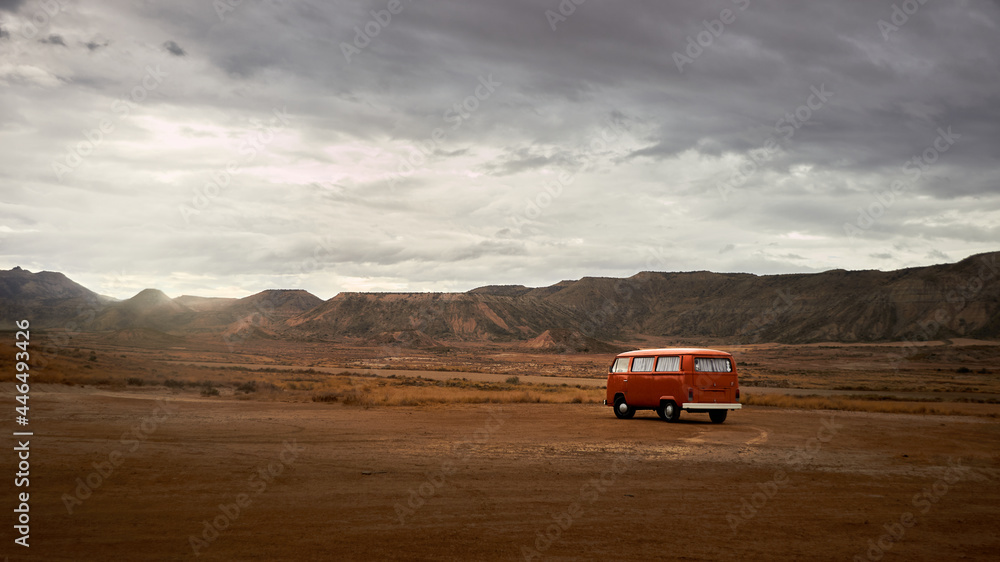 orange camper van parked in the desert for the night, red sand and mountains in the background with a cloudy sky