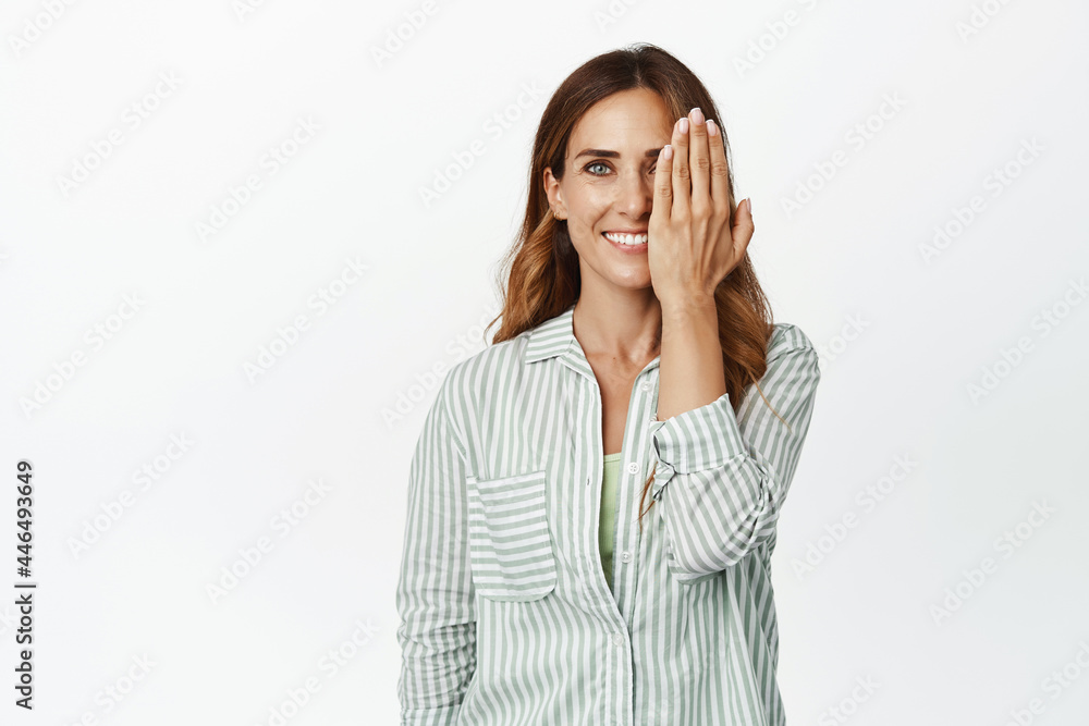 Portrait of smiling brunette woman, middle aged businesswoman in blouse, cover half side of face with hand, looking one eye with happy facial expression, white background Portrait of smiling brunette woman, middle aged businesswoman in blouse, cover half side of face with hand, looking one eye with happy facial expression, white background