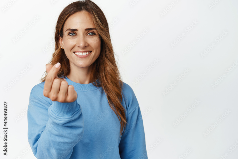 Portrait of beautiful and happy woman smiling, showing finger heart gesture, standing in casual clothes against white background Portrait of beautiful and happy woman smiling, showing finger heart gesture, standing in casual clothes against white background