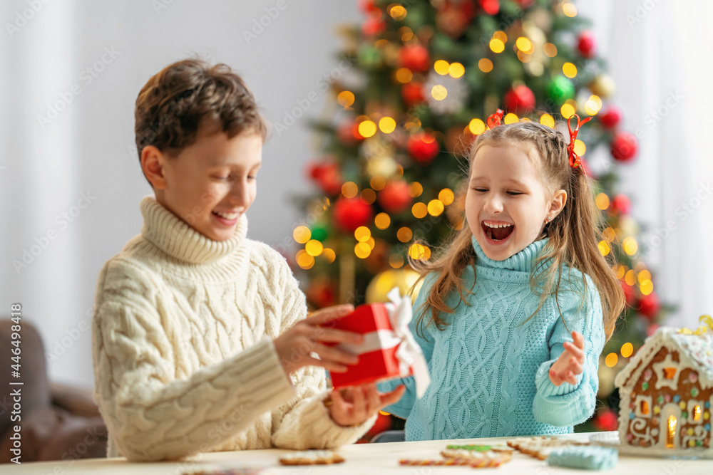 boy gives gift box to little girl. baby is happy and laughs when receiving gift. beautifully decorated room with Christmas tree, gingerbread house on table, boy and girl look at each other and laugh. boy gives gift box to little girl. baby is happy and laughs when receiving gift. beautifully decorated room with Christmas tree, gingerbread house on table, boy and girl look at each other and laugh.