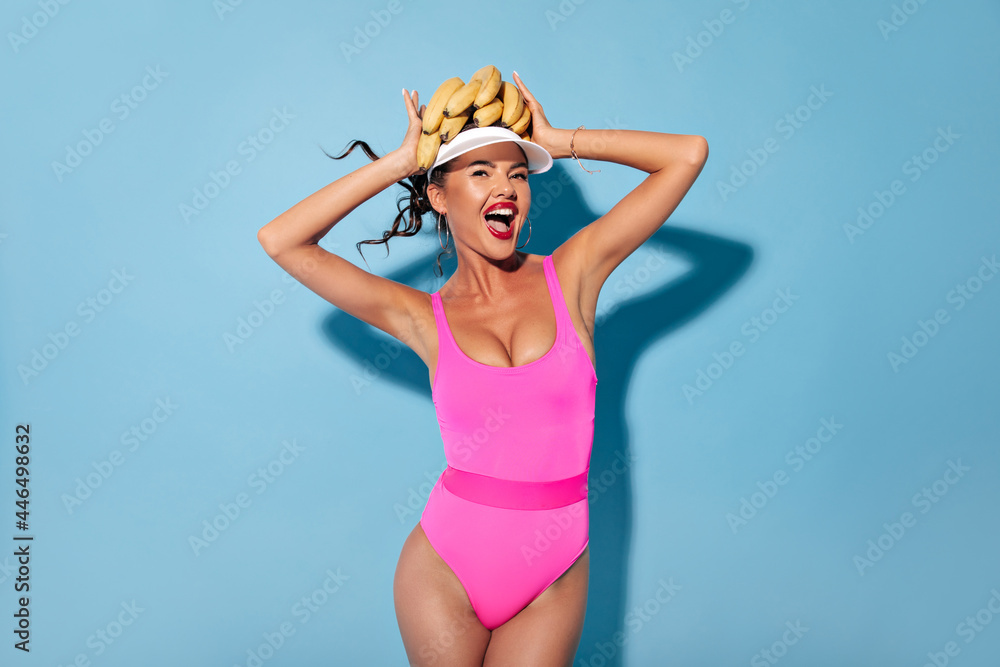 Modern cool young woman with curly hair, silver earrings and bright lipstick in pink swimming suit and white cap posing with bananas..