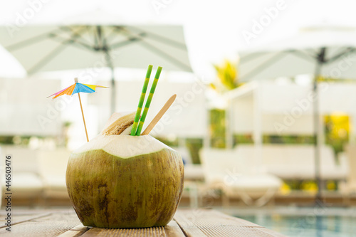 Fresh green coconut drink with paper strawand umbrella  standing on wooden board near pool  water  tropical  resort background  with copy space  .Vacation exotic spa resort travel destinations concept