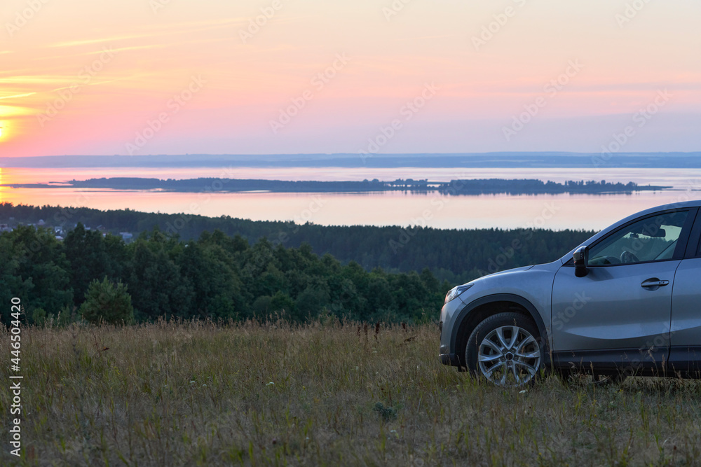 A quiet summer evening. A fragment of a car standing on a high river bank. The sun is setting over the river. The rays of the setting sun are reflected on the water.