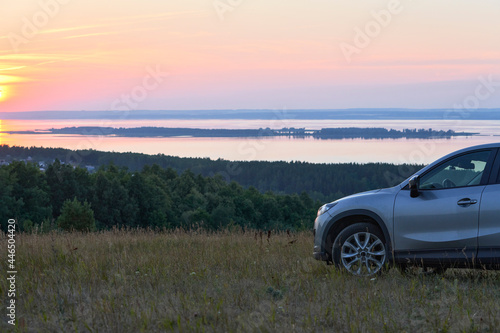 A quiet summer evening. A fragment of a car standing on a high river bank. The sun is setting over the river. The rays of the setting sun are reflected on the water.