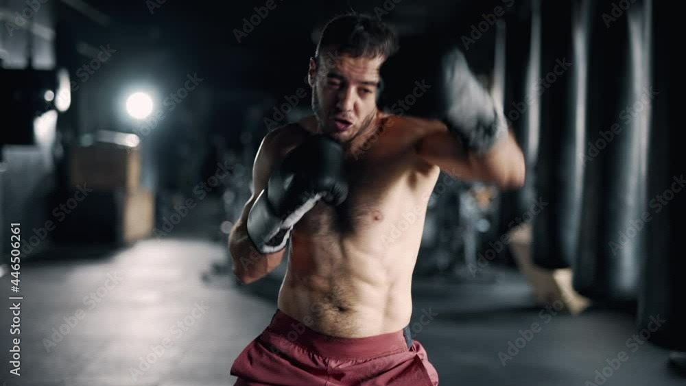 A muscular sportsman is boxing in the dark gym. Self confident boxer is looking at the camera and training indoors. Maximum focus and strength. Concept of lifestyle health and sports. Close up. A muscular sportsman is boxing in the dark gym. Self confident boxer is looking at the camera and training indoors. Maximum focus and strength. Concept of lifestyle health and sports. Close up.