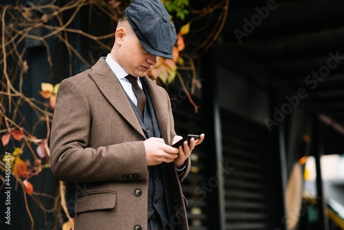 A man posing in the image of an English retro gangster of the 1920s dressed in Peaky blinders style near old brick wall.