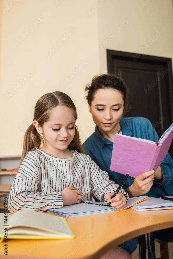 young teacher reading book near girl writing dictation in montessori school