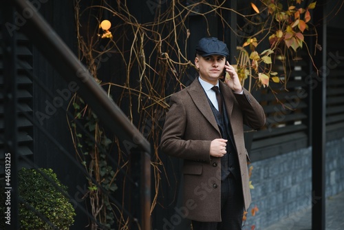 A man posing in the image of an English retro gangster of the 1920s dressed in Peaky blinders style near old brick wall.