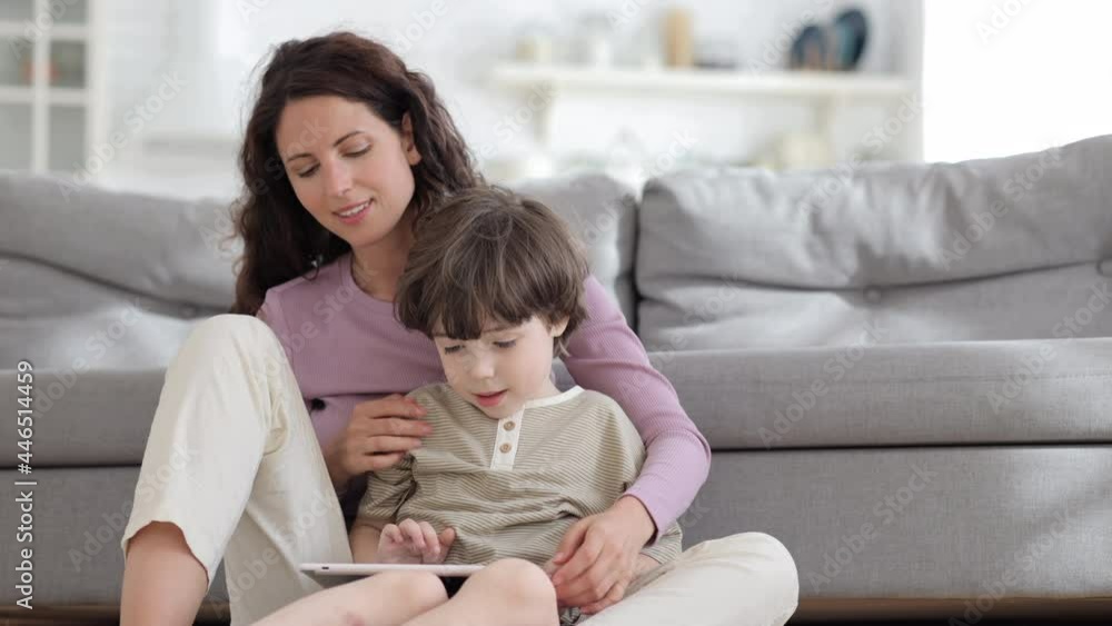 Young single mother or baby sitter relax with small boy and digital tablet: mom and kid sitting cuddling with touchscreen computer in hands smiling watch funny video or cartoon online at home together Young single mother or baby sitter relax with small boy and digital tablet: mom and kid sitting cuddling with touchscreen computer in hands smiling watch funny video or cartoon online at home together