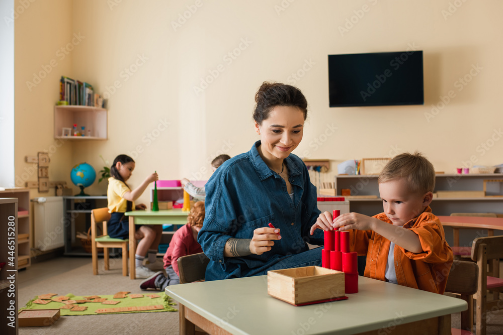 young teacher smiling near boy making tower from wooden cylinders near kids on blurred background young teacher smiling near boy making tower from wooden cylinders near kids on blurred background