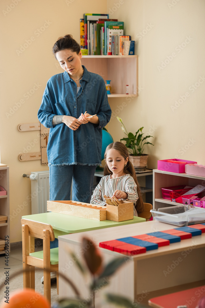 Obraz premium young teacher standing near girl playing educational game in montessori school