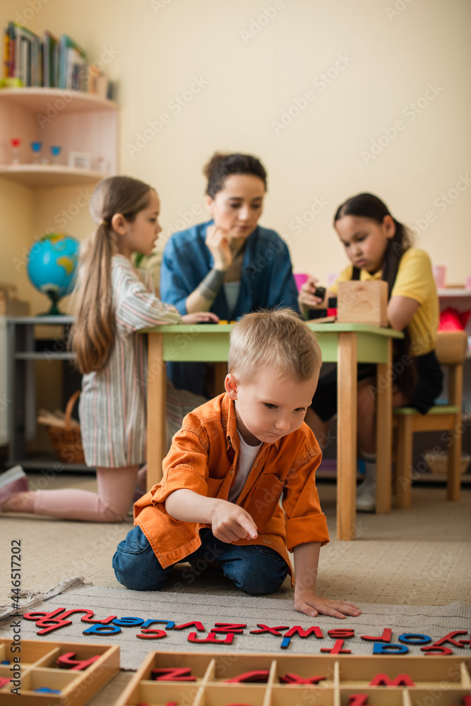 Fototapeta premium boy on floor pointing at wooden letters near teacher and interracial kids on blurred background