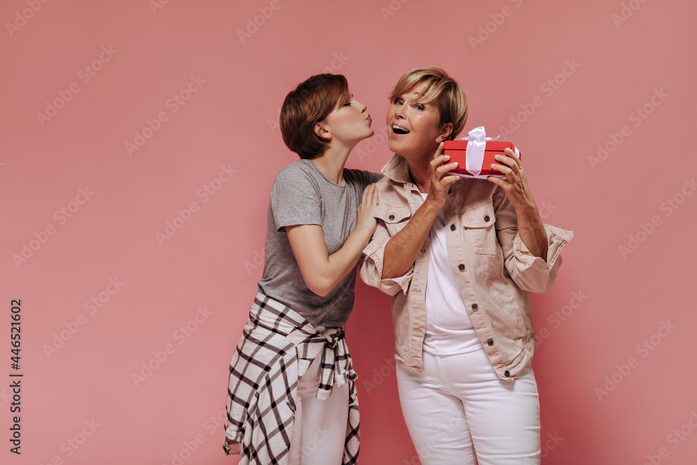 Beautiful brunette girl in shirt and pants kissing her grandmother in cheek on pink backdrop. Blonde woman posing with red gift box and cool lady.. Beautiful brunette girl in shirt and pants kissing her grandmother in cheek on pink backdrop. Blonde woman posing with red gift box and cool lady..