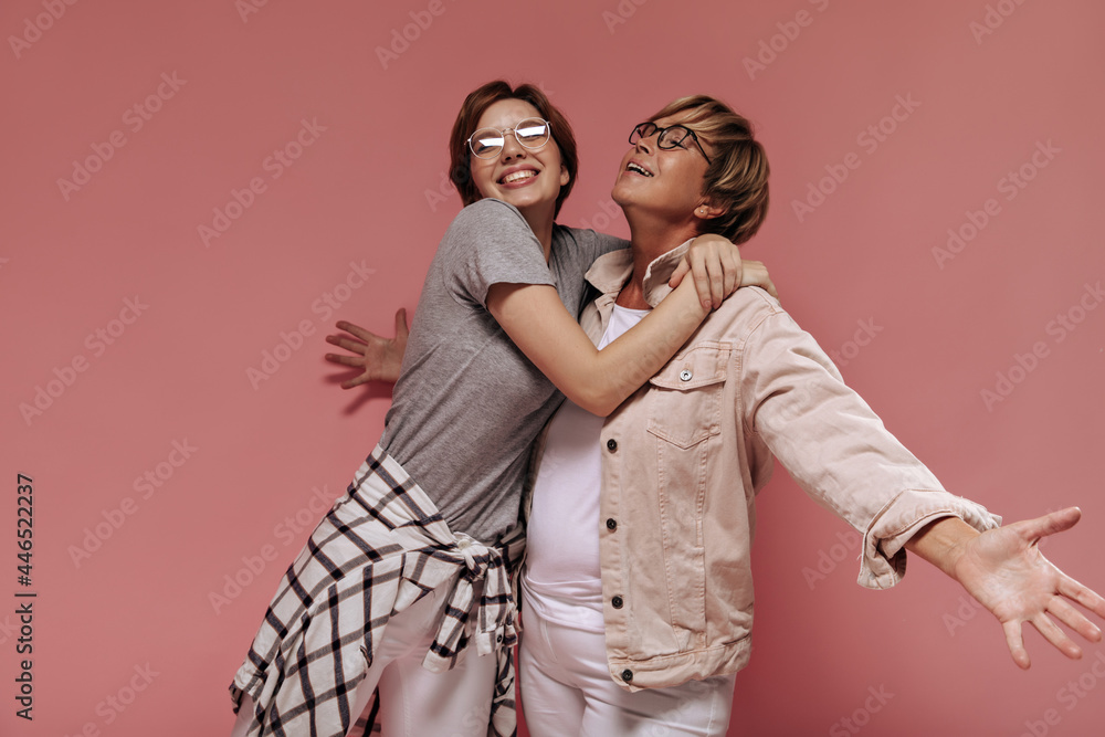 Positive two short haired women with modern glasses in fashionable light clothes hugging and smiling on pink background.. Positive two short haired women with modern glasses in fashionable light clothes hugging and smiling on pink background..