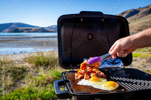 A man cooks bacon and eggs on a portable bbq beside the seaside on a sunny winter day travelling in New Zealand