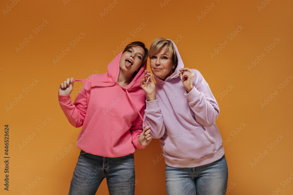 Brunette lady showing tongue in pink clothes and posing with blonde woman in lilac hoodies on orange isolated background.. Brunette lady showing tongue in pink clothes and posing with blonde woman in lilac hoodies on orange isolated background..