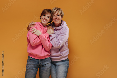 Charming two ladies with lovely smiles and short cool hairstyle in modern pink sweatshirts and trendy jeans hugging on isolated backdrop..
