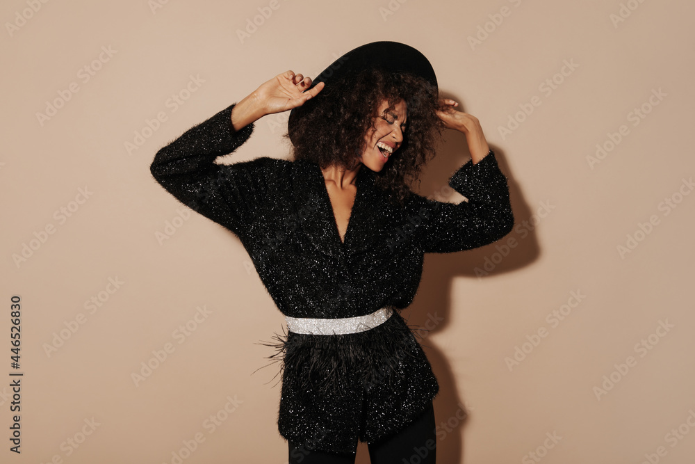 Fototapeta premium Cheerful woman with brunette wavy hairstyle in hat and fashionable black sweater with light belt laughing on beige background..