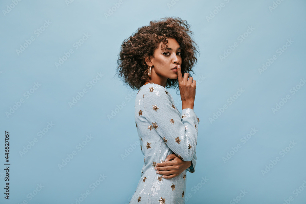 Fashionable girl with curly hair and modern makeup in cool blue clothes and gold earrings looking into camera on isolated background..