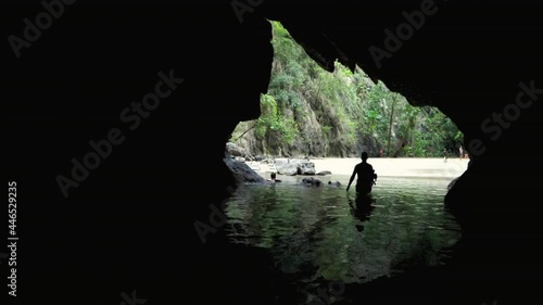 Hand held shot of male snorkeler entering emerald cave in Koh Muk Thailand. A silhouette exits the ocean water and walks through a dark cave towards the white sand beach with karst mountain background photo