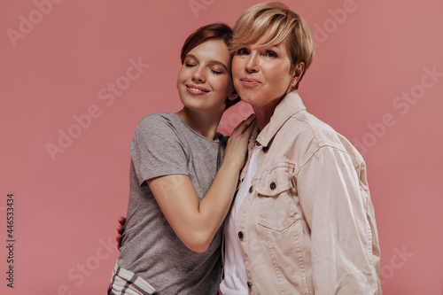 Blonde short haired woman in beige jacket looking into camera and hugging with young girl in grey t-shirt with closed eyes on pink backdrop..