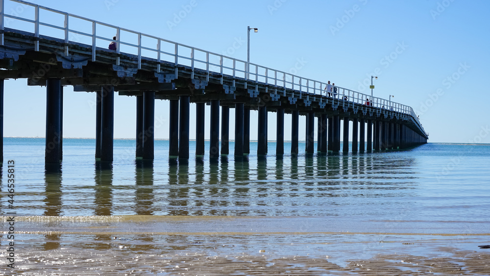 Fototapeta premium Looking up at long wooden pier, with blue sky, calm water, gentle waves