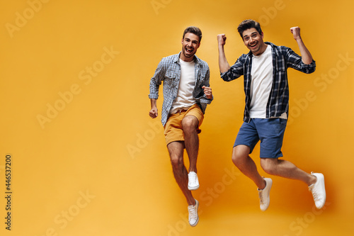 Charming handsome young men in colorful shorts and checkered shirts have fun, rejoice and jump on orange background.