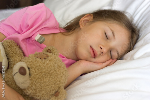 A cute little girl with blond hair in a pink T-shirt lies asleep in the bed, measures the temperature and hugs a teddy bear. photo
