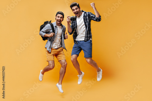 Cool happy men in shorts and checkered shirts jump and rejoice on orange background. Happy guys hold retro camera and backpack.