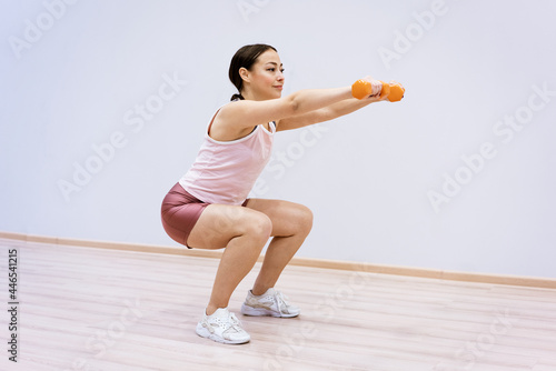 A woman of Caucasian ethnicity in sportswear squats with dumbbells in her hand against the background of a light wall. Home sports concept