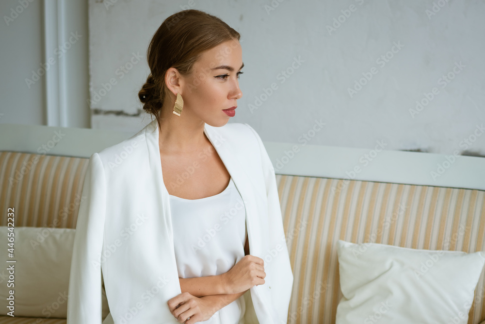 Young business woman of Caucasian ethnicity is sitting on the sofa in white business clothes with beautiful makeup and hair. Portrait of a confident beautiful girl Young business woman of Caucasian ethnicity is sitting on the sofa in white business clothes with beautiful makeup and hair. Portrait of a confident beautiful girl