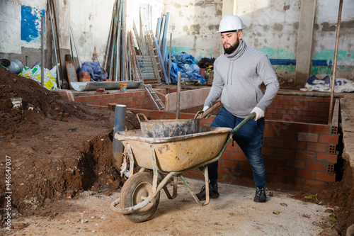 Young worker carrying bucket with cement mortar on small cart in building under construction