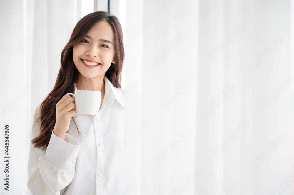 Young asian beautiful woman clothing white pyjamas and drink coffee beside window at morning in bedroom. She feeling happy fresh and smile relax Young asian beautiful woman clothing white pyjamas and drink coffee beside window at morning in bedroom. She feeling happy fresh and smile relax