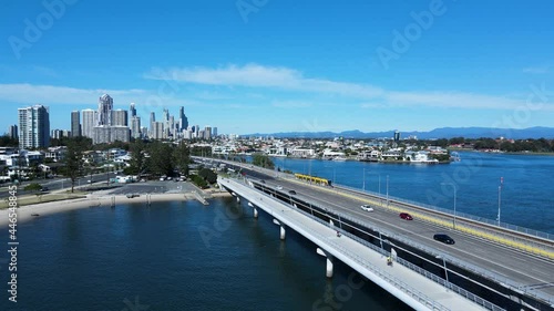 Fast modern electric tram travelling across a urban city bridge leading to a towering skyline backdrop. High panoramic drone view. photo