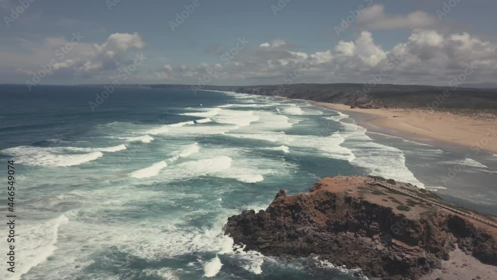 Waves of blue Atlantic Ocean reaching shore of sandy beach in Portugal. Sunny day with beautiful coastline and clouds at sky. Waves of blue Atlantic Ocean reaching shore of sandy beach in Portugal. Sunny day with beautiful coastline and clouds at sky.