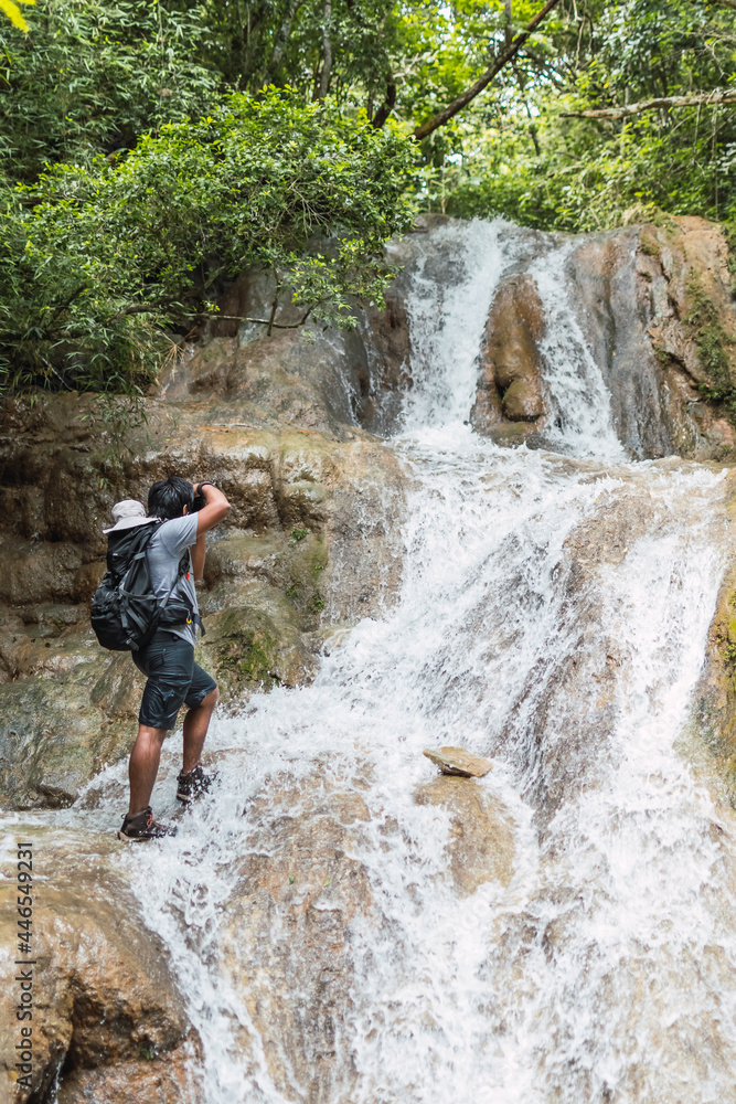 Young man with backpack in waterfall in mountain taking photos and hikign 
