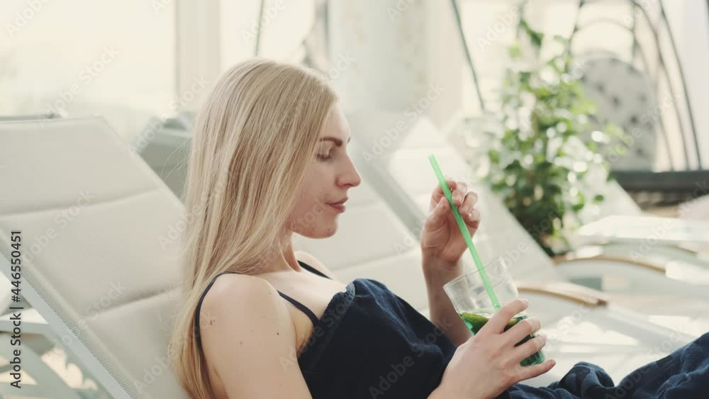 Medium shot of blonde young woman drinking cooling mint drink from a straw. She lying on a deck chair and relaxing. Medium shot of blonde young woman drinking cooling mint drink from a straw. She lying on a deck chair and relaxing.