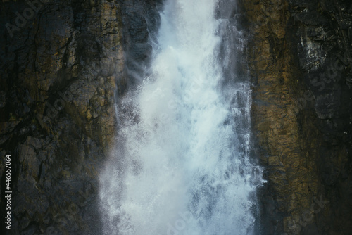 Atmospheric minimal landscape with vertical big waterfall on rock mountain wall. Powerful large waterfall in dark gorge. Nature background of high vertical turbulent falling water stream on wet rocks.