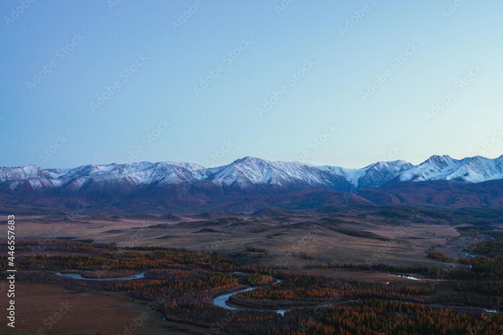 Awesome landscape with mountain river serpentine in valley among hills and forest in autumn colors with view to great snowy mountain range in sunset. High snow-covered mountains and autumn valley.