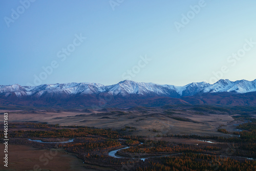 Awesome landscape with mountain river serpentine in valley among hills and forest in autumn colors with view to great snowy mountain range in sunset. High snow-covered mountains and autumn valley. © Daniil