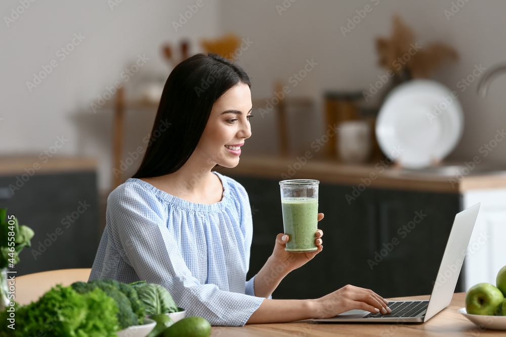 Young woman with glass of smoothie using laptop in kitchen