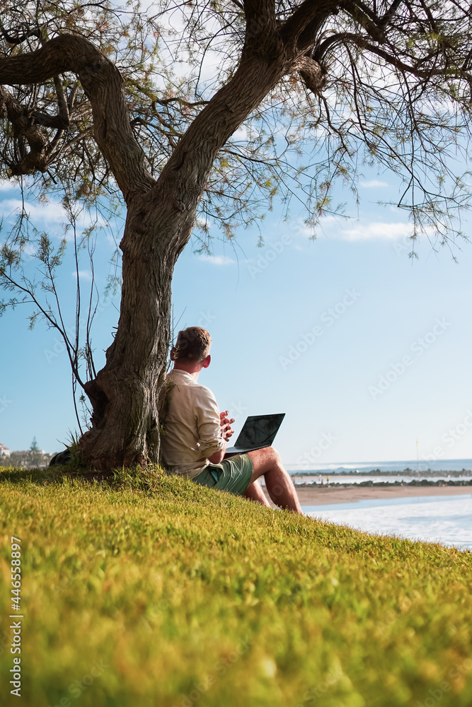 remote working business man sitting relaxed and lean against a tree with laptop and sea view on green grass at the beach to work 