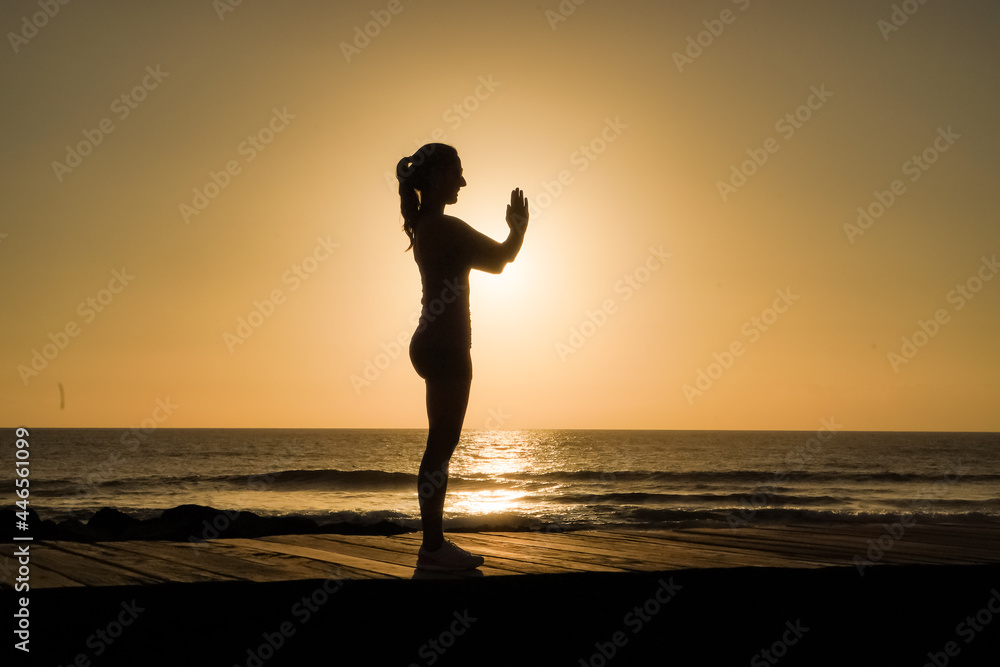 silhouette of a sportive female in namaste pose practicing yoga & meditation on beach boardwalk during sunset or sunrise 